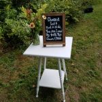 A Welcoming Sign On A White Stand Amidst Greenery Instructing Wedding Guests To Choose A Seat, Not A Side, As They'Re Loved By Both The Bride And Groom.