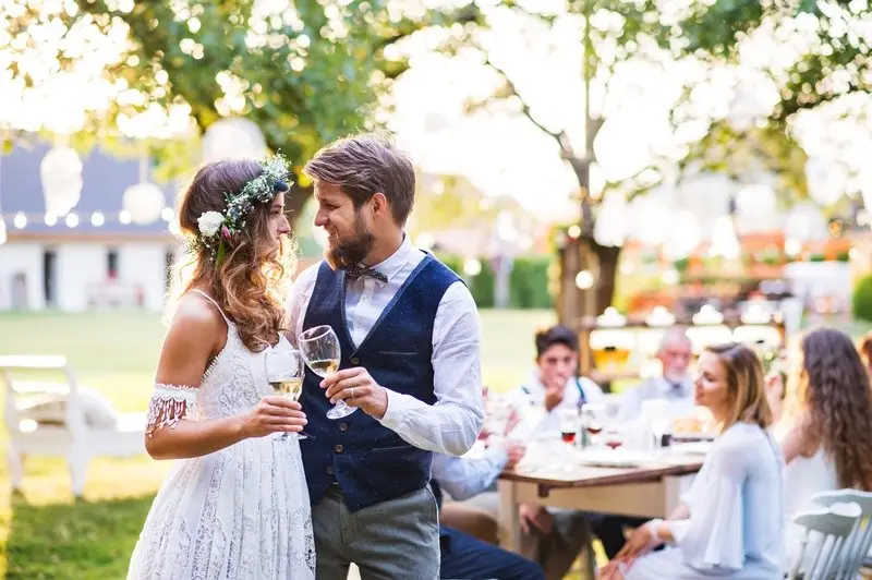 A Couple In Wedding Attire Is Toasting With Wine Glasses At An Outdoor Celebration With Guests In The Background.