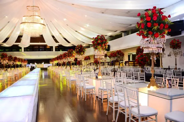 An Elegant Banquet Hall With White Chairs, A Central Aisle, And Tables Adorned With Red Floral Centerpieces And Chandeliers.
