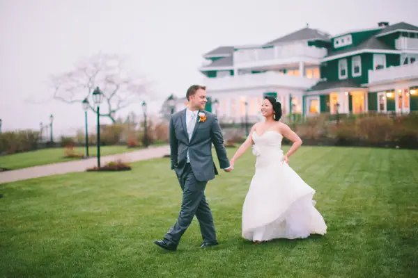 A Bride And Groom Are Walking Hand In Hand On A Grassy Lawn With A House In The Background.